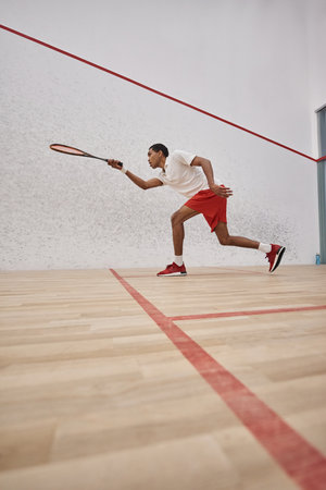 energetic african american sportsman in shorts holding racquet and playing squash inside of courtの写真素材