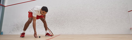 african american man in red shorts holding racquet and picking up squash ball in court, bannerの写真素材
