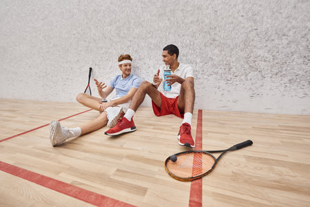 cheerful interracial sportsmen with squash racquets and bottle with water sitting on floor in courtの写真素材