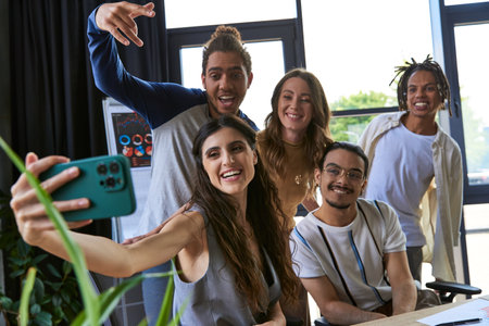 happy woman taking photo on smartphone with excited multicultural team in coworking officeの写真素材