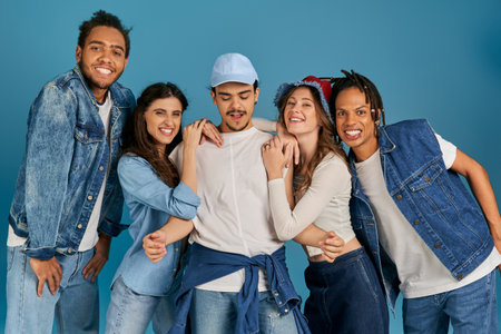 cheerful and trendy multicultural friends posing near young man in cap on blue, modern fashion