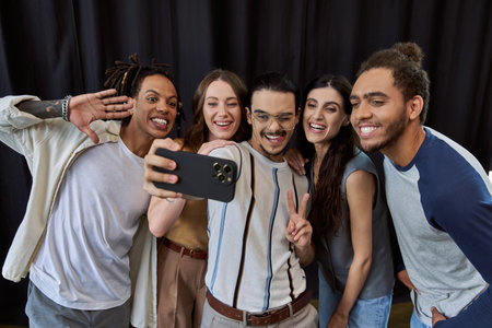happy man in eyeglasses showing victory sign and taking photo on smartphone with multiethnic teamの写真素材
