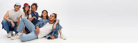 trendy african man in denim clothes lying down near happy multiethnic friends posing on grey, banner