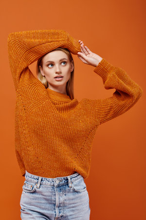young blonde woman in orange knitted sweater posing with arms above head on orange, winter vibesの写真素材