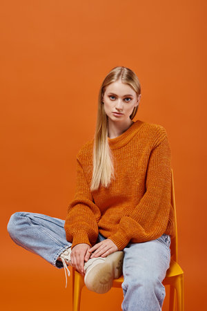 trendy blonde woman in warm winter sweater and jeans sitting on chair looking at camera on orangeの写真素材