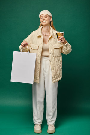 happy woman in stylish winter clothing with shopping bag, takeaway drink and credit card on greenの写真素材