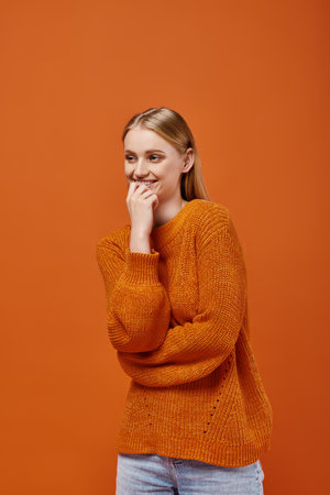smiling blonde woman in bright knitted sweater smiling and looking away on orange, winter emotionsの写真素材