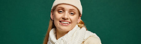 trendy woman in white knitted scarf and beanie hat smiling at camera on green, winter style bannerの写真素材