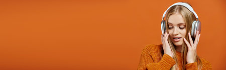 blonde woman in orange knitted sweater and headphones listening music on bright backdrop, bannerの写真素材