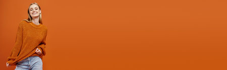 happy blonde woman stretching her bright sweater while posing on orange backdrop, bannerの写真素材