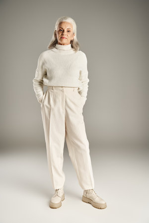 attractive and grey haired middle aged woman in white winter outfit standing with hands in pocketsの写真素材