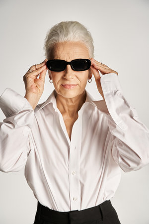 stylish middle aged woman with grey hair wearing sunglasses and standing in elegant attire on greyの写真素材