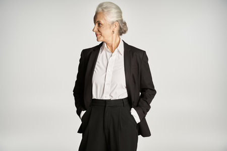 smiling middle aged businesswoman in formal wear standing with hands in pockets on grey backgroundの写真素材