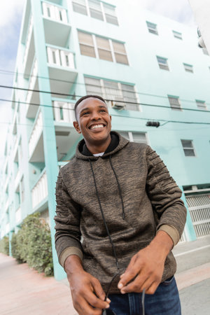 low angle view of happy african american man in hoodie holding stylish sunglasses while standing near blue building in Miamiの写真素材