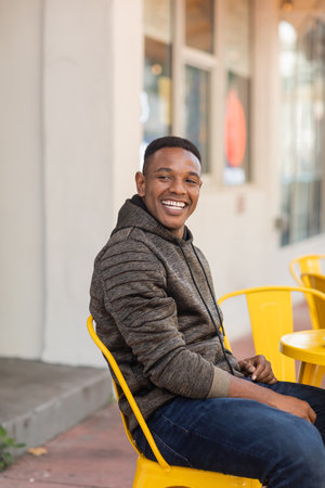 cheerful african american man in hoodie and jeans sitting at bistro table in outdoor cafe
