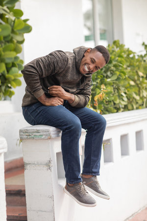 happy african american man in hoodie and jeans holding sunglasses in teeth while sitting outdoorsの写真素材
