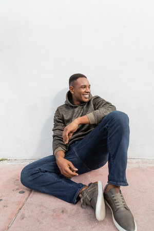 cheerful african american man in jeans and hoodie smiling while sitting near white wallの写真素材