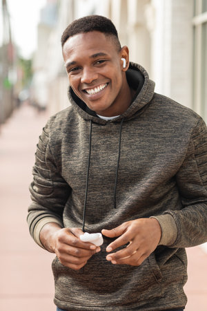 cheerful african american man in hoodie and wireless earphone listening music and holding case on street in Miamiの写真素材