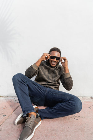 smiling african american man in stylish sunglasses sitting near white wall