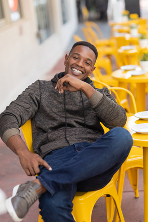 positive african american man in hoodie and jeans sitting at yellow bistro table in outdoor cafeの写真素材