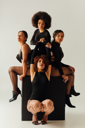 attractive african american women in black alluring outfits posing on black cube, fashion conceptの写真素材