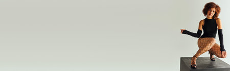 appealing young african american woman in sexy black attire squatting on black cube, fashion, bannerの写真素材