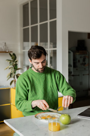 bearded man in green jumper holding cutlery near breakfast in modern kitchen at homeの写真素材