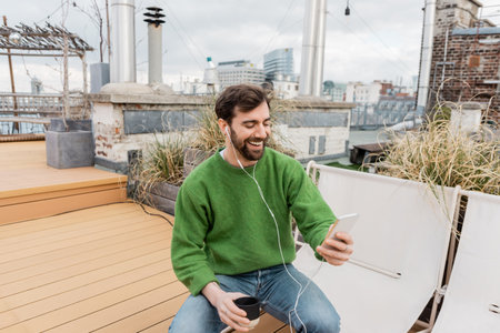 Cheerful man in earphones holding cup of coffee while spending time on rooftop terrace in Viennaの写真素材