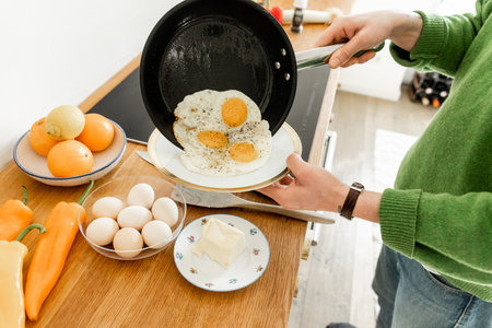 Cropped view of man holding pan with fried eggs near plate while cooking breakfast in morning