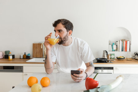 Bearded and tattooed man in white t-shirt drinking fresh orange juice and using smartphone