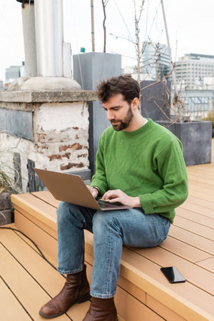 handsome freelancer using laptop near smartphone while working on rooftop in Vienna, Austria