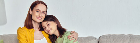 happy woman embracing teenage daughter and looking at camera in living room, horizontal banner
