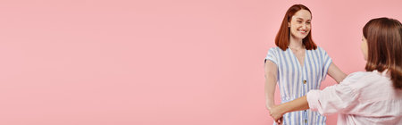 joyful and trendy woman with teenage daughter holding hands on pink backdrop, horizontal bannerの写真素材