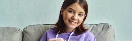 cheerful teenage girl sitting on couch with smartphone and looking at camera in living room, banner