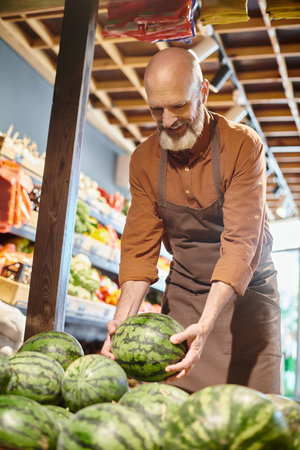 joyful bearded mature seller smiling happily and picking fresh watermelon at grocery storeの写真素材