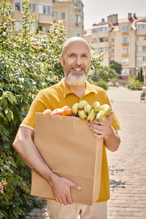 cheerful mature male customer posing outside with paper bag full of fruits and smiling at cameraの写真素材
