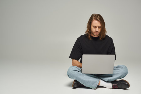 handsome man with long hair and beard sitting with crossed legs on floor and using laptop on greyの写真素材