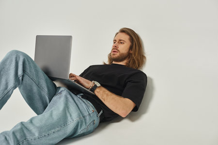 handsome man with long hair and beard leaning on white wall and using laptop on grey backdropの写真素材