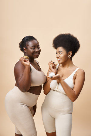happy african american plus size women in lingerie holding cosmetic cream on beige, skincareの写真素材