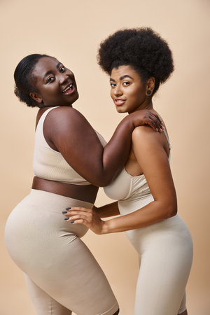 sensual and smiling plus size african american women posing in lingerie on beige, curvy beautyの写真素材