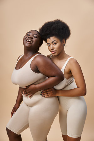 joyful plus size african american women in underwear looking at camera on beige, body positivityの写真素材