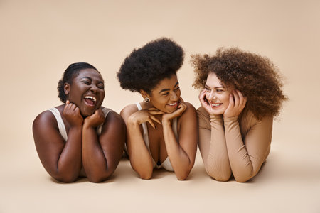 smiling multiethnic body positive women in lingerie lying down and smiling at each other on beigeの写真素材