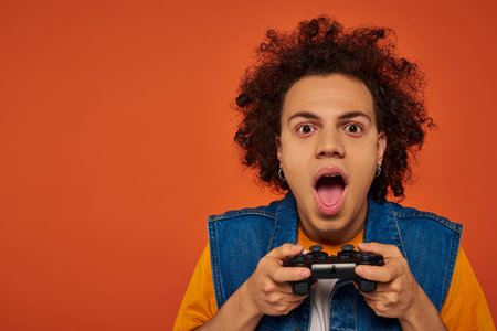attractive african american man in casual attire emotionally playing video games on orange backdrop