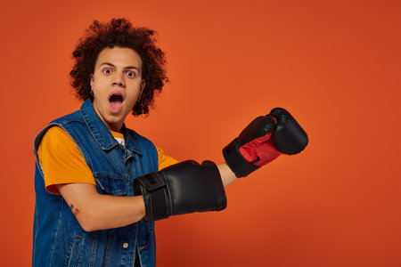 surprised sporty african american male model posing lively in boxing gloves on orange backgroundの写真素材