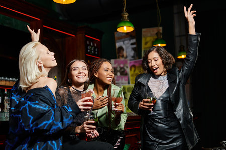 charming and happy multiethnic girlfriends dancing with cocktail glasses in bar, glamorous partyの写真素材