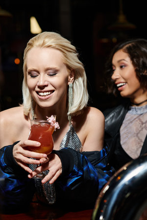delighted blonde woman with glass of delicious cocktail near multicultural girlfriend in barの写真素材