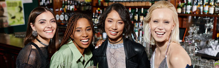 joyful and stylish multiethnic girlfriends with cocktail glasses looking at camera in bar, bannerの写真素材