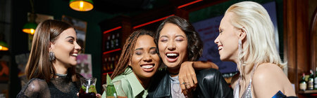 cheerful multiracial girlfriends with cocktail glasses embracing and laughing in bar, bannerの写真素材