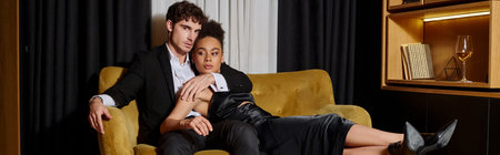 multicultural couple in black evening attire posing together while sitting on velvet sofa, bannerの写真素材