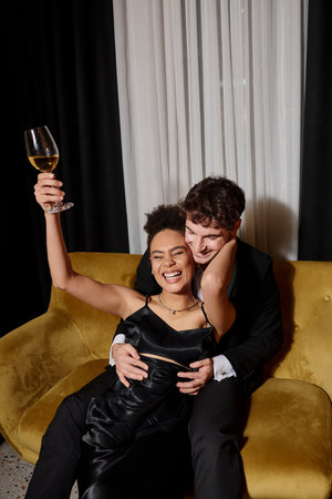 happy african american woman holding glass of wine and laughing near man in suit on velvet sofaの写真素材